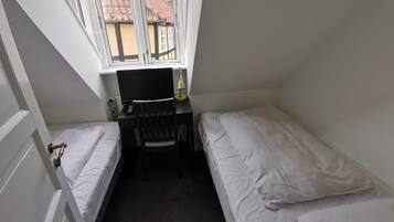 Una habitación pequeña y ordenada con dos camas individuales, un escritorio con silla y ventana con vistas a un edificio de tejado a la vista.