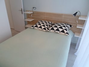 A bed with a light-colored mattress, two patterned pillows, a bedside table with a lamp, and a wall-mounted shelf with a book and a phone.