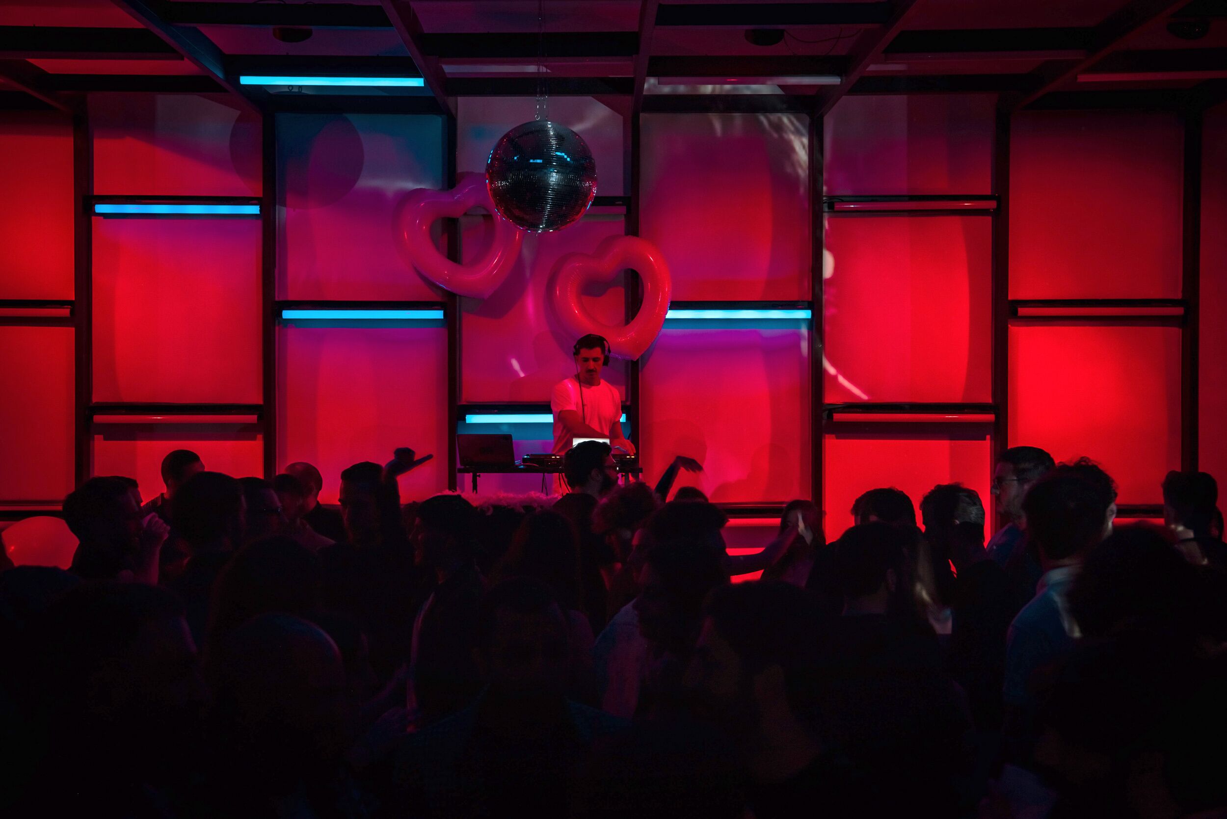 A DJ booth with a disco ball and heart-shaped decorations, surrounded by a crowd in a dimly lit room.