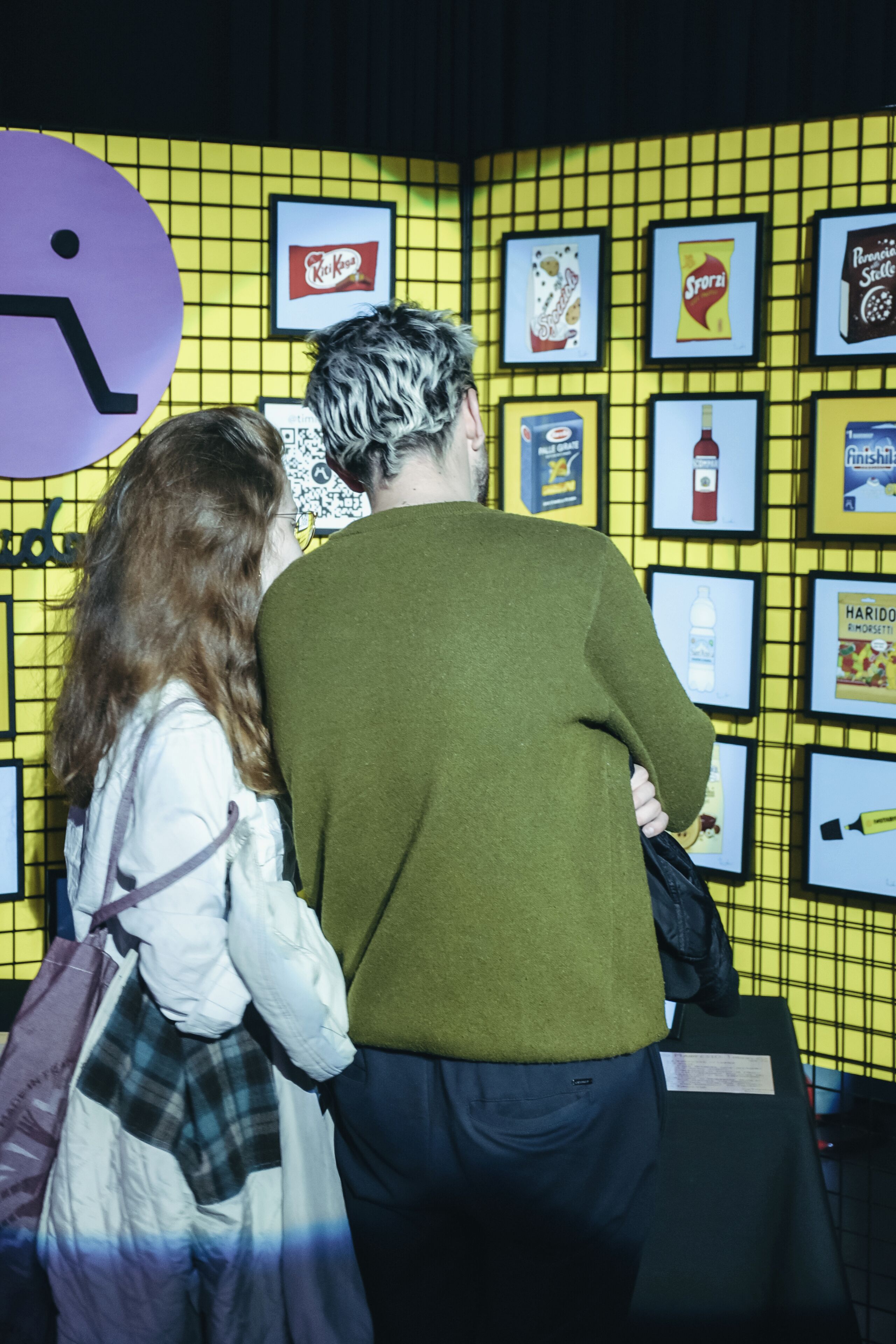 Two people examining framed items on a yellow grid wall.