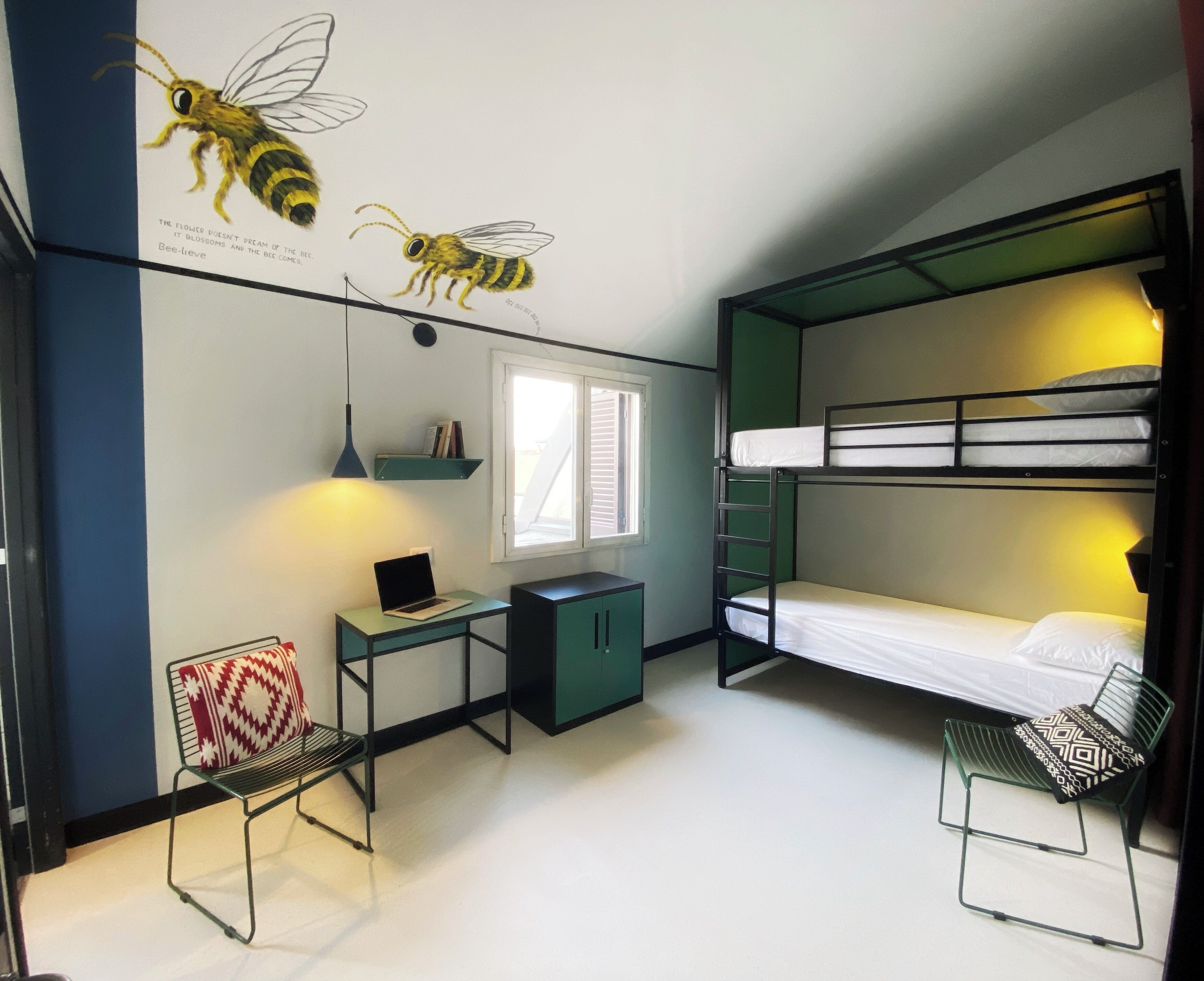A dormitory room with bunk beds, a desk with a laptop, a chair, a small cabinet, and a window with blinds.