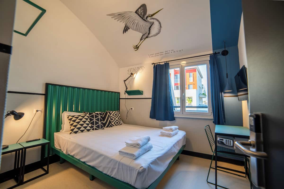 A neatly made bed with white linens and towels, a green headboard, a desk with a chair, a wall-mounted TV, and a window with curtains.