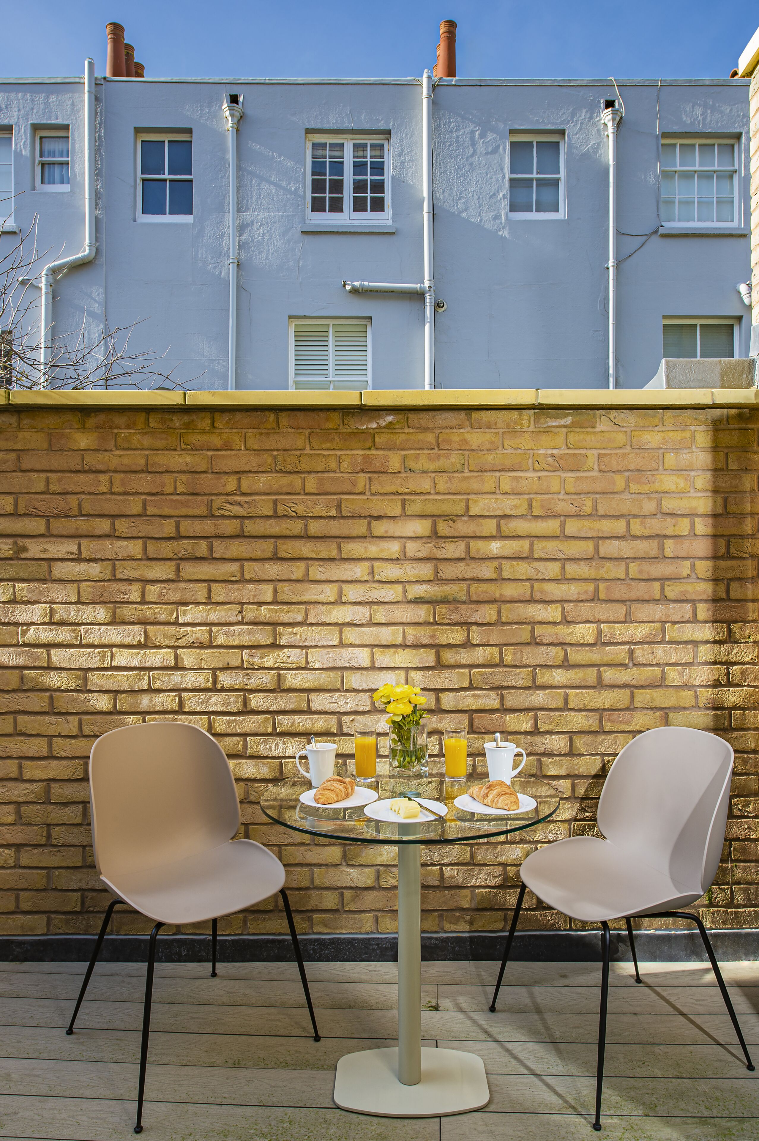 A patio with two chairs and a table, a brick wall, and a building in the background.