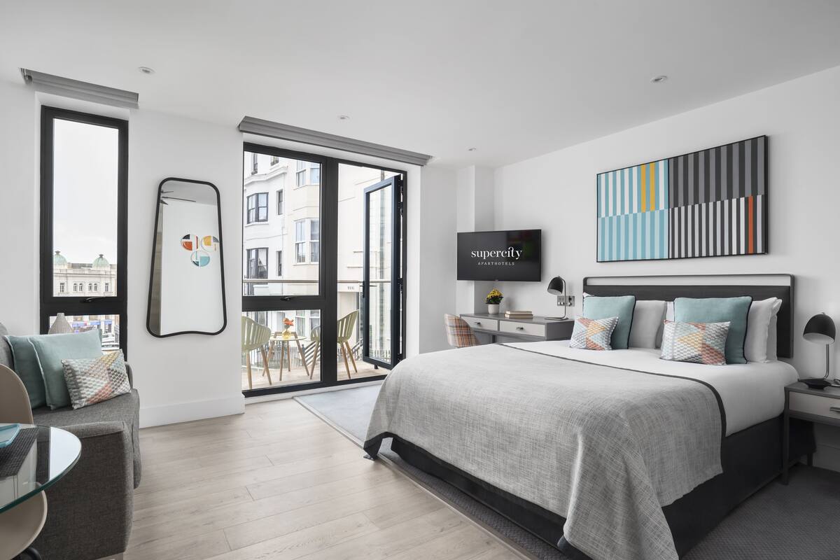 Een moderne slaapkamer met een groot bed, een televisie, een bureau en een balkon met uitzicht op gebouwen.