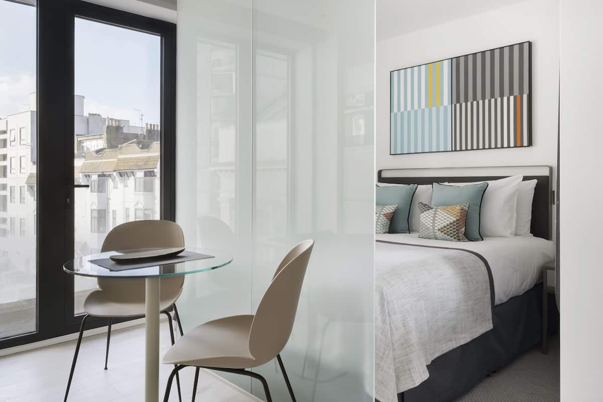 Een moderne hotelkamer met een glazen scheiding, een bed met gestreepte kussens, een klein rond tafeltje met een stoel, en uitzicht over de stad.