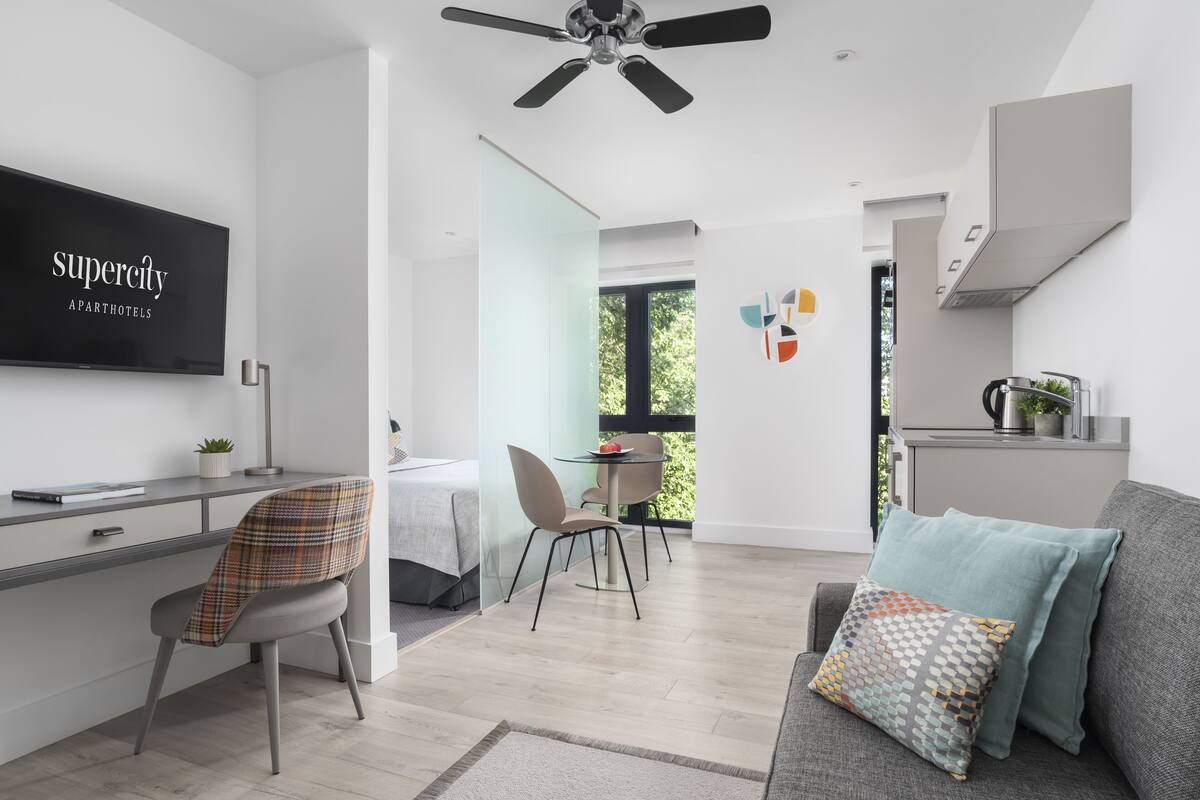 Een moderne woonkamer met een grijze bank, een televisiescherm waarop 'supercity apartments' staat, een eethoek met een tafel en stoelen, en een keuken met een fornuis en kastjes.