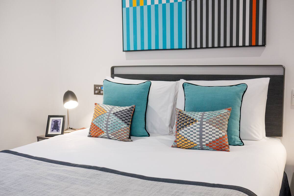 Een net opgemaakt bed met een witte deken en teal-kleurige kussens, een ingelijst foto op het nachtkastje, en een modern abstract schilderij aan de muur.