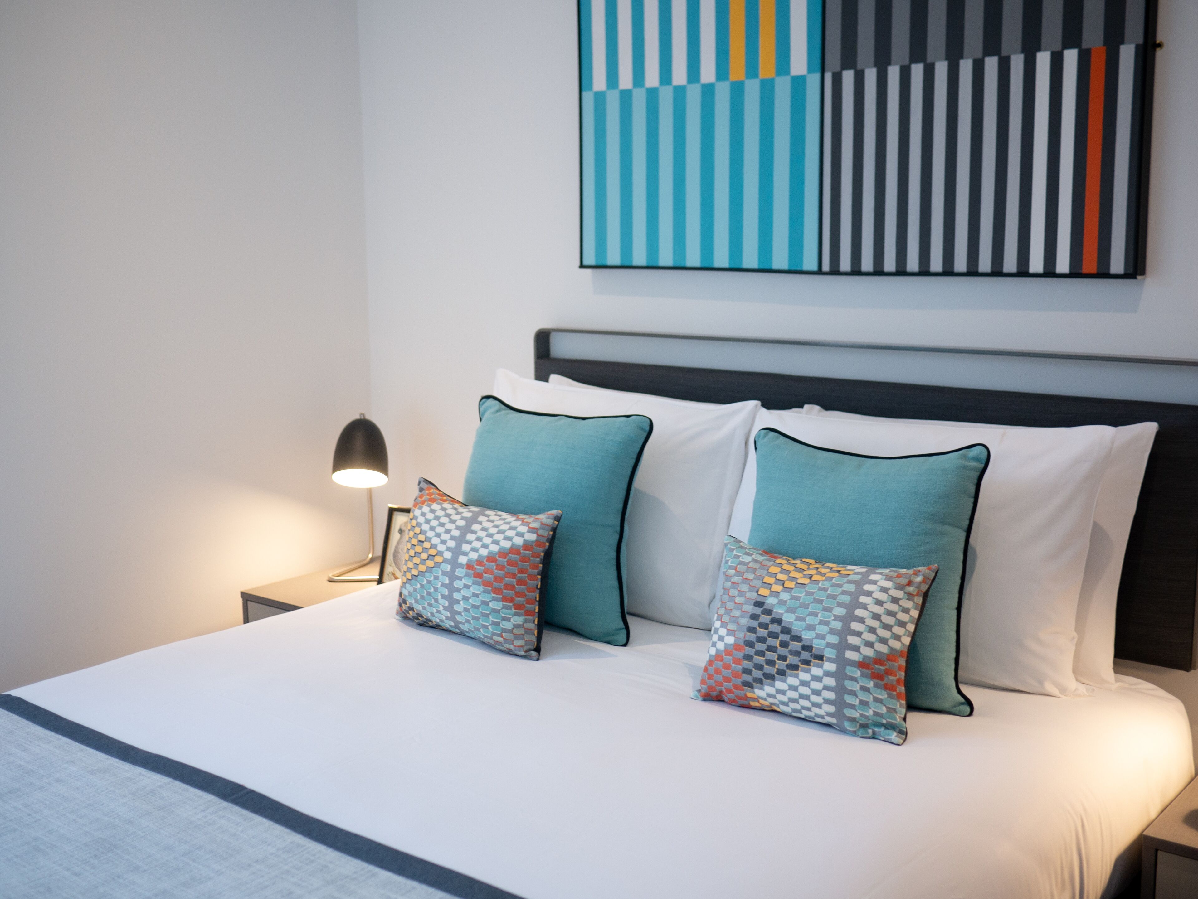 A neatly made bed with two patterned pillows, a bedside lamp, and a modern abstract painting on the wall.