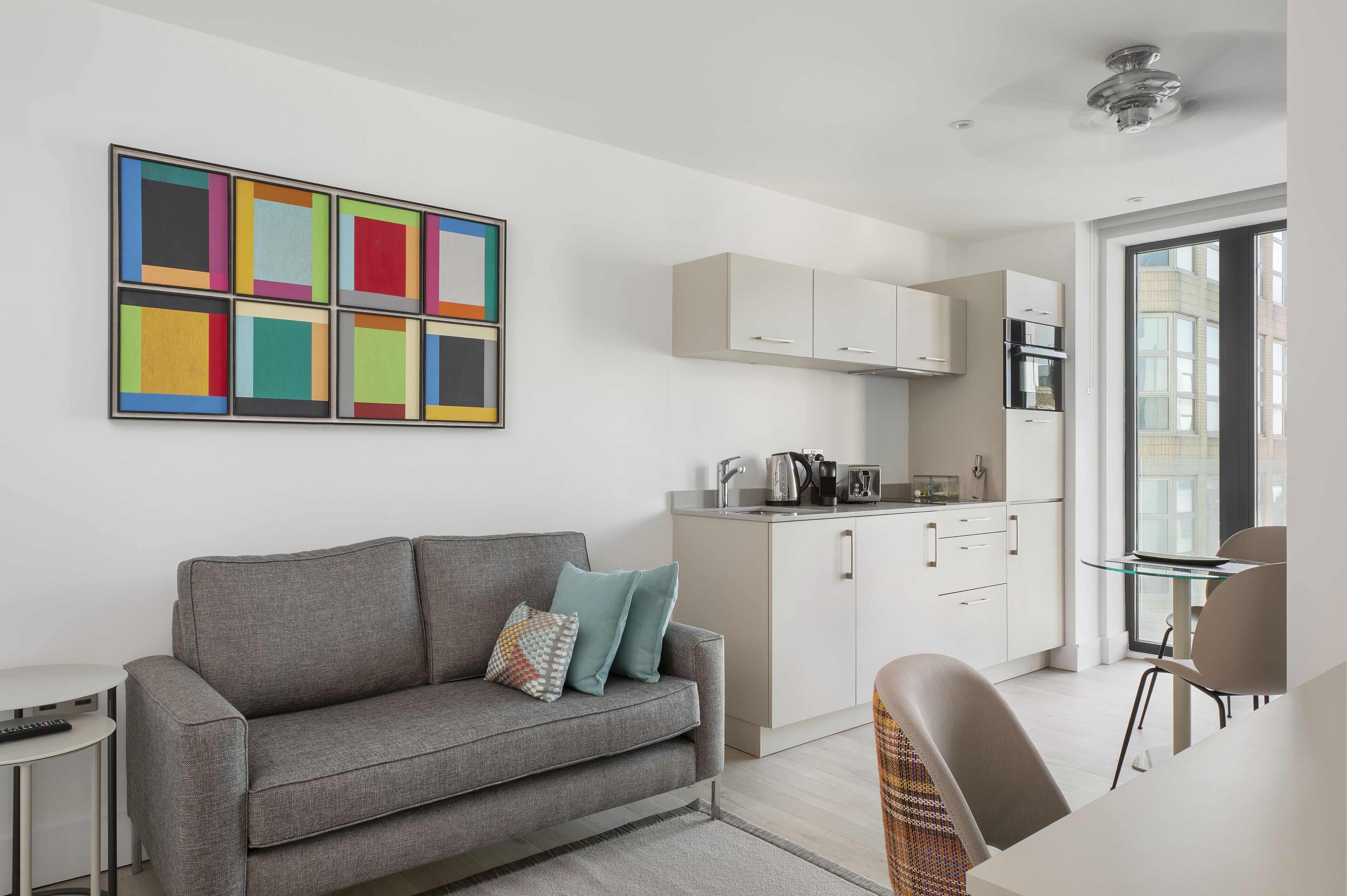 A modern apartment with a grey sofa, a colorful abstract painting, a kitchen with white cabinets, and a view through a large window.