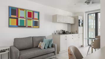 Een modern appartement met een grijze bank, een kleurrijk abstract schilderij, een keuken met witte kasten en een uitzicht door een groot raam.