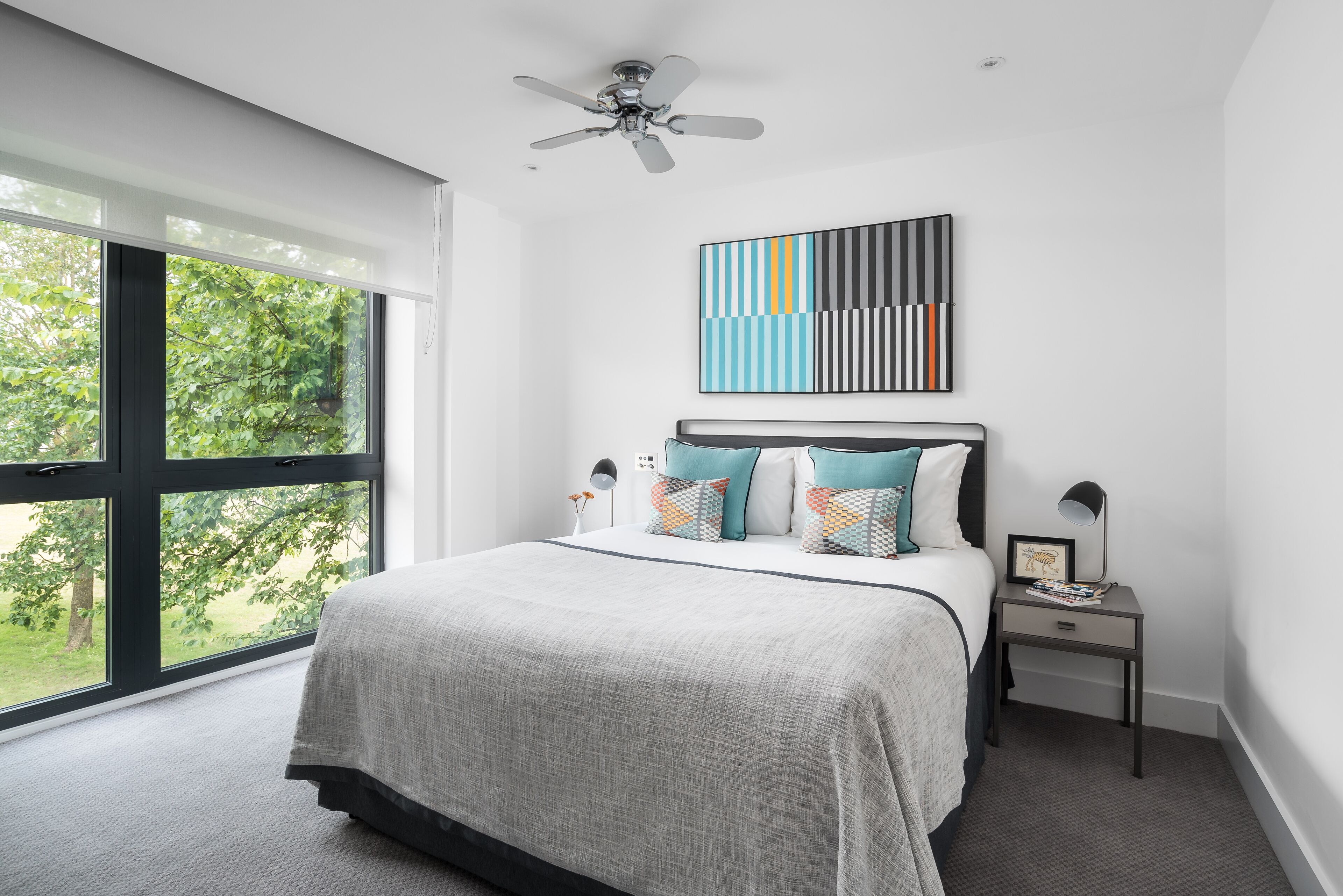 Une chambre moderne avec une grande fenêtre, un lit agrémenté d’oreillers à motifs, une table de chevet avec une lampe et un ventilateur de plafond.