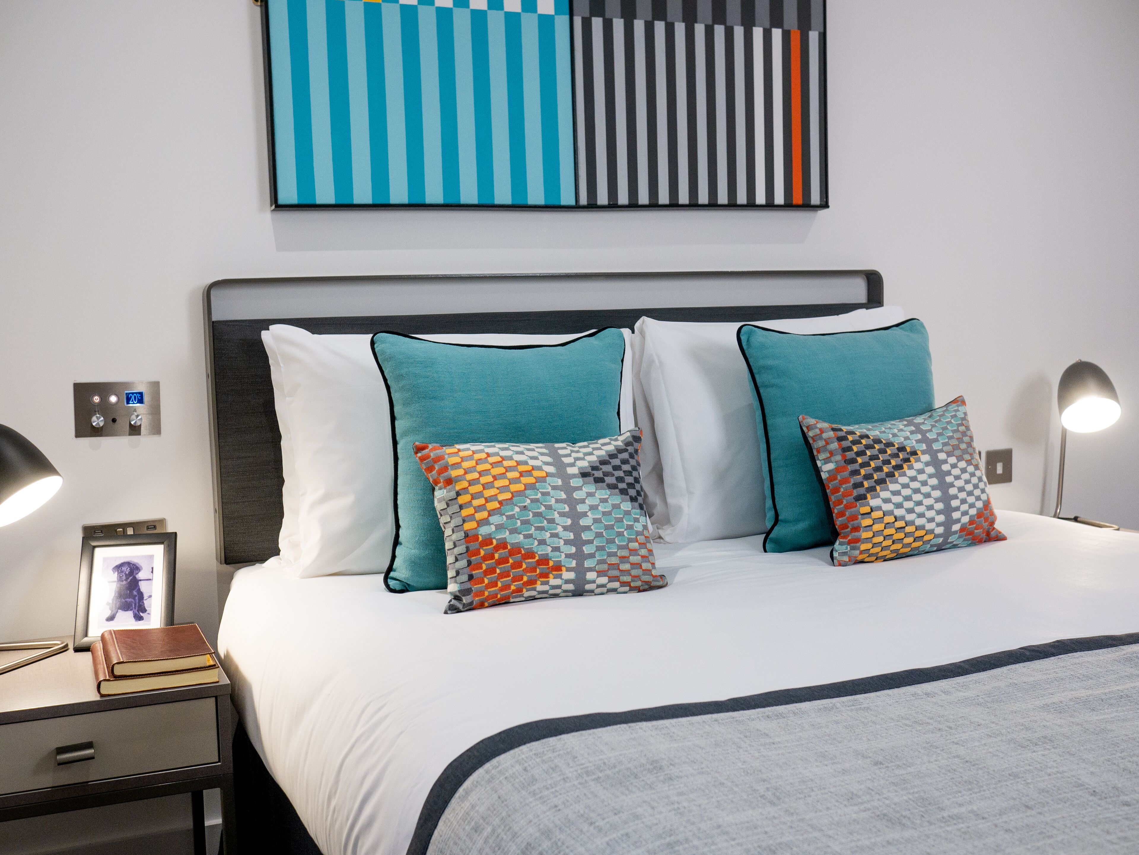 A neatly made bed with white linens, teal and patterned pillows, and a modern headboard. A bedside table with a lamp and a framed photo is visible.
