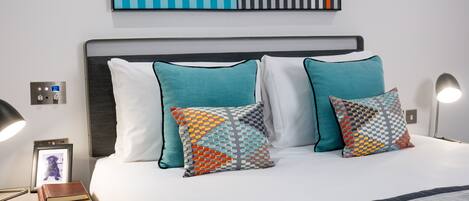 A neatly made bed with white linens, teal and patterned pillows, and a modern headboard. A bedside table with a lamp and a framed photo is visible.