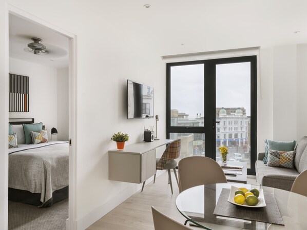 A modern apartment with a bed, a desk, a dining area, and a large window with a city view. - Q Square by Supercity Aparthotels (Brighton)