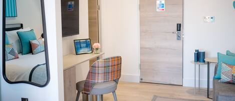 A modern hotel room with a bed, a desk, a chair, a mirror, and a door.