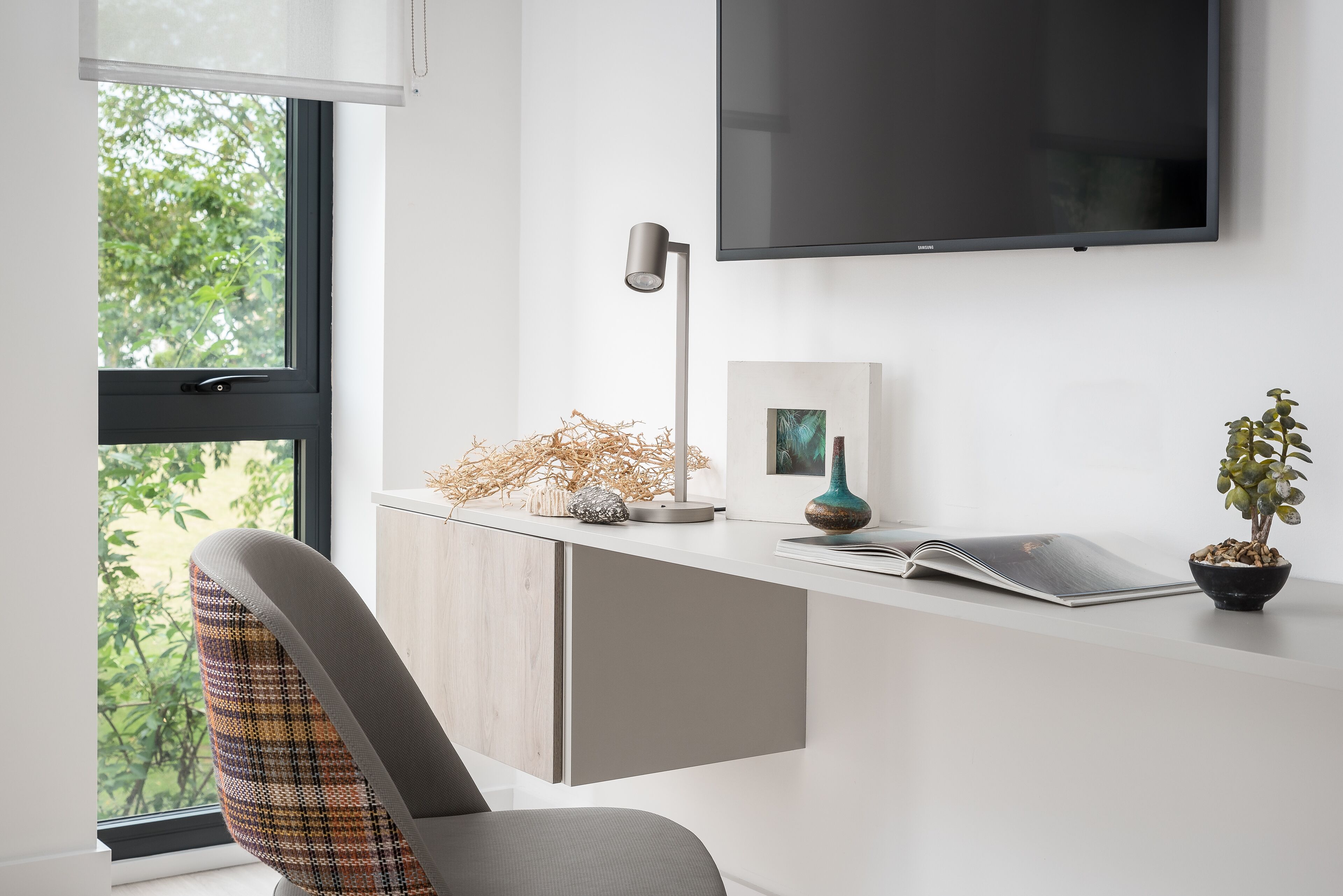 A modern office space with a desk, chair, lamp, TV, and a window with a view of greenery.