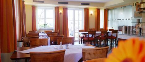 A dining area with multiple tables and chairs, large windows with curtains, and a kitchen area in the background.