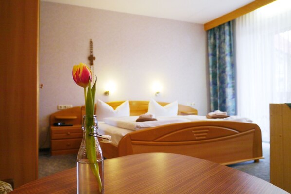 A hotel room with a bed, bedside tables, a wooden nightstand, a vase with a single flower, and a window with curtains. - Landgasthof Zum Hirschen (Kemnath)