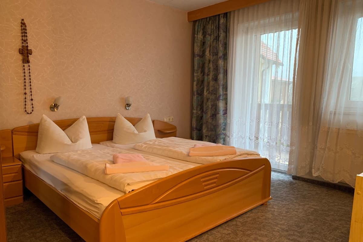 Habitación de hotel con cama de madera, ropa de cama blanca, una mesita de noche, una lámpara fijada a la pared y una ventana con cortinas traslúcidas.