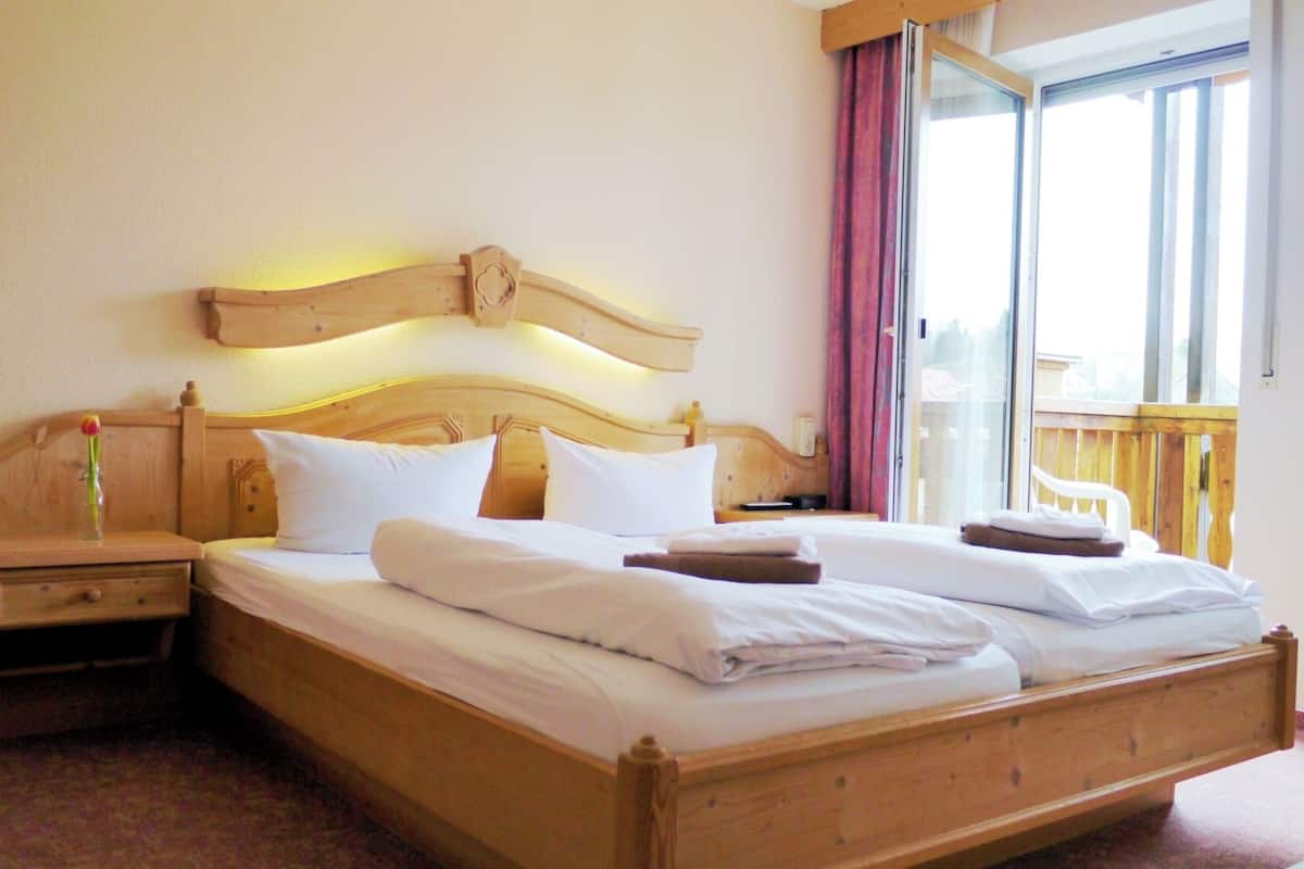 Cama doble con cabecera de madera y lámparas en la cabecera, mesita de noche con un jarrón y ventana con vistas a una terraza de madera.