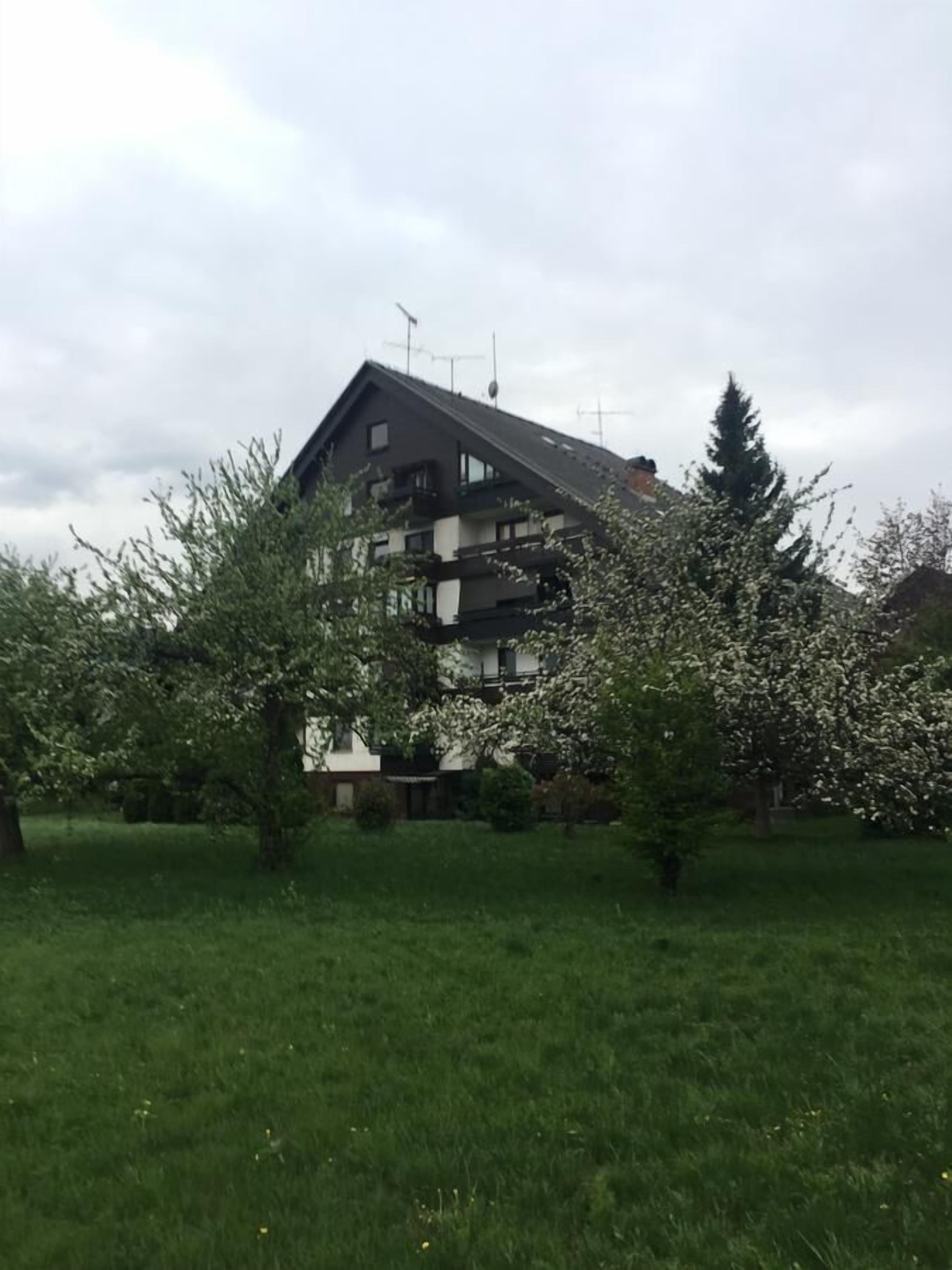 Un immeuble résidentiel à plusieurs étages, doté d’un toit en pente et entouré d’une zone verdoyante avec des arbres.