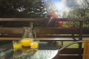 Una mesa de vidrio con una jarrón y dos vasos de jugo de naranja, sobre un balcón con baranda de madera y vistas a árboles y cielo.