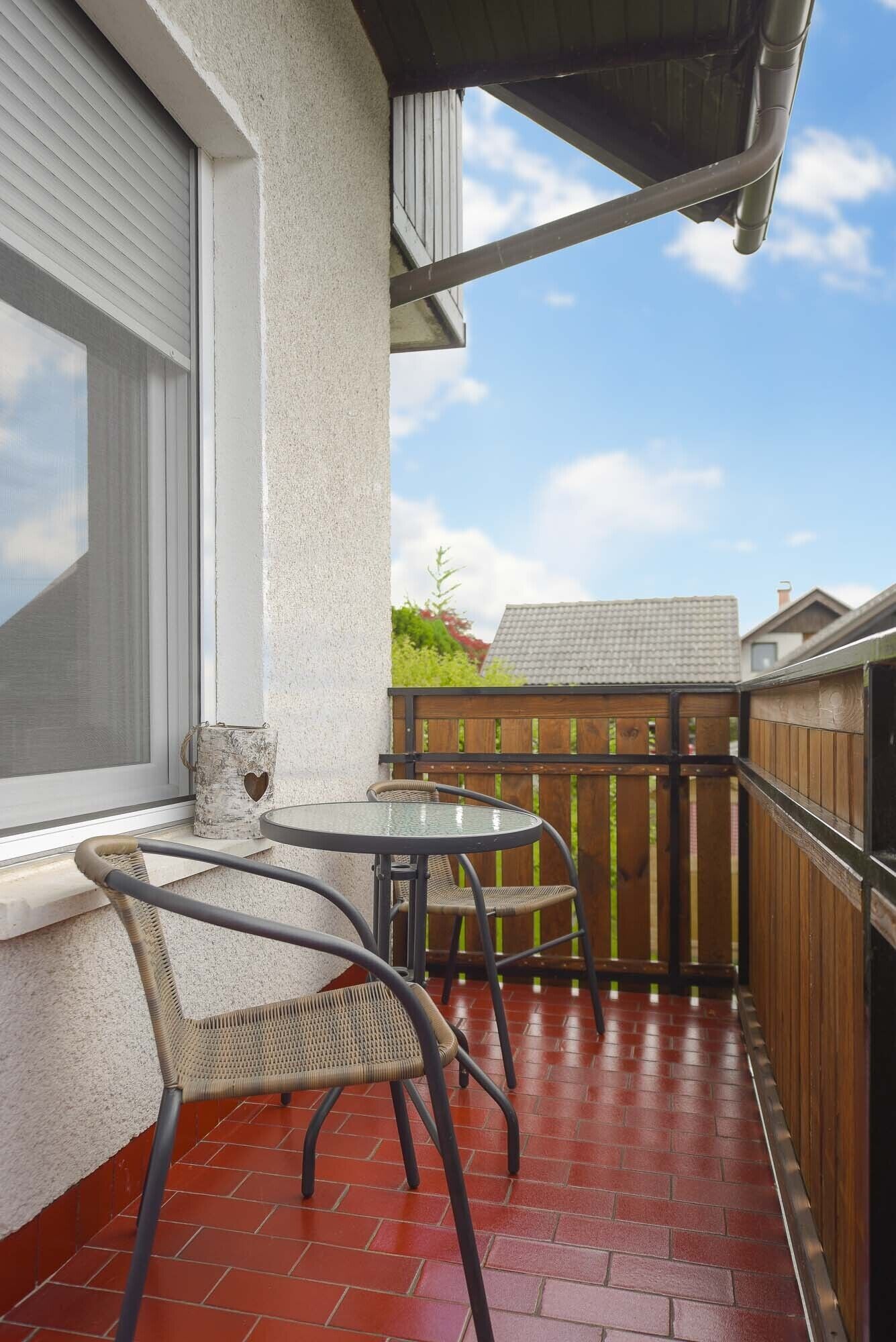 A small patio with a round table and two wicker chairs, a wooden fence, and a window with blinds.