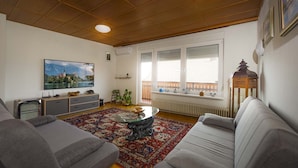 Uma sala de estar com sofá, mesa de centro, televisão e uma janela grande com persianas.