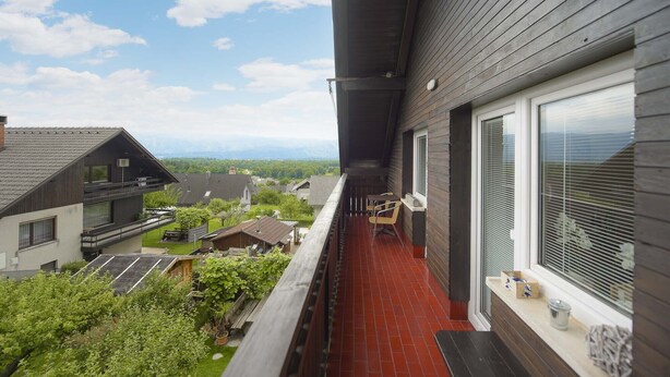 Uma varanda com piso de telhas vermelhas, com vista para uma área residencial com casas e uma cordilheira ao longe.