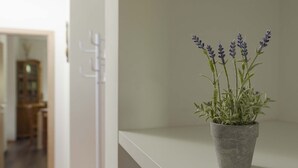 Uma planta de lavanda em vaso sobre uma estante branca num quarto, com uma porta e um armário de madeira ao fundo.