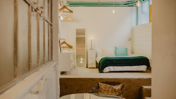 Une chambre d’hôtel de taille réduite, comprenant un lit, une petite table et une chaise. On y trouve également une armoire avec un miroir et une tringle pour suspendre les vêtements.
