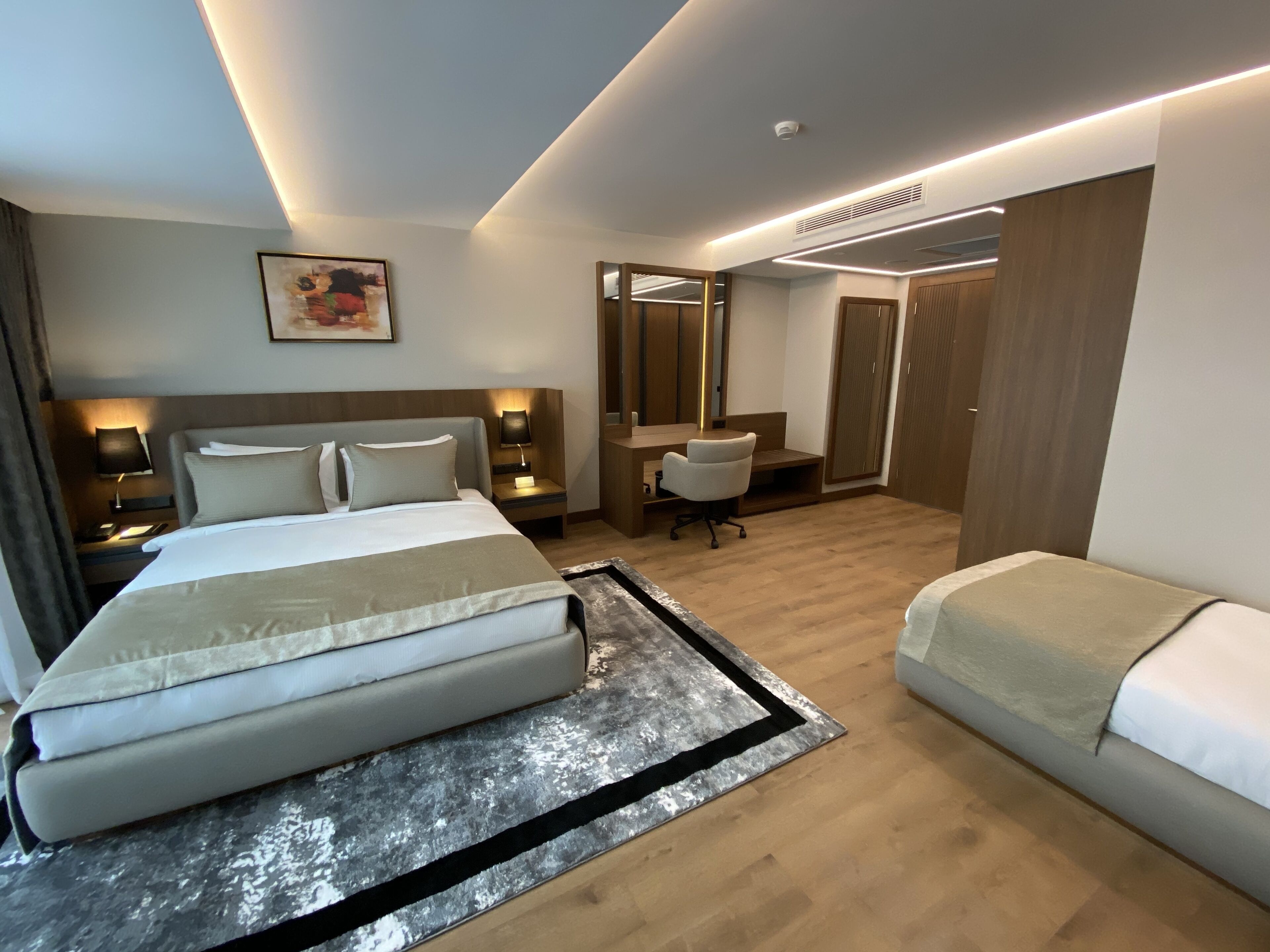 A modern hotel room with a large bed, bedside tables with lamps, a desk with a chair, and a wardrobe.