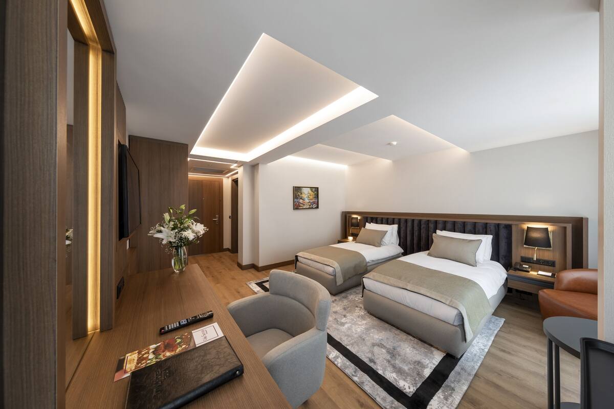 A modern hotel room with a large bed, a smaller bed, a desk, and a chair. There is a painting on the wall and a vase with flowers on the desk.