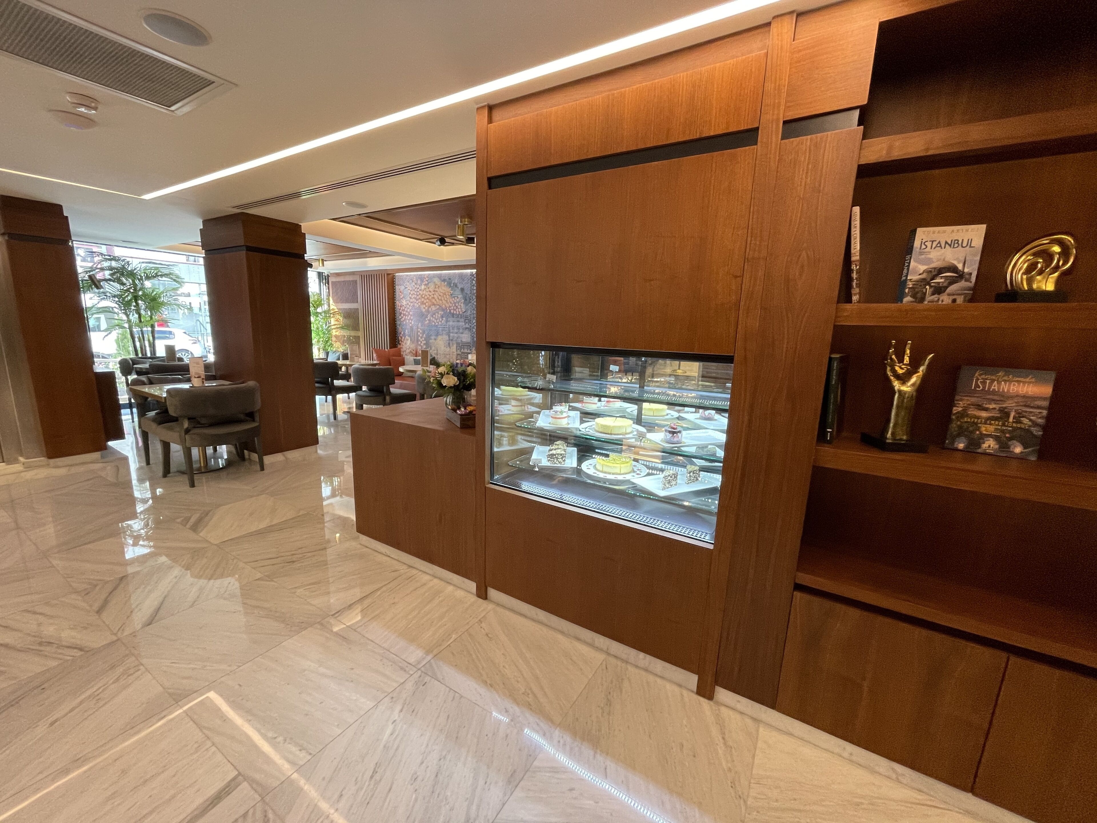 Un vestíbulo de hotel con suelo de mármol, una mesa de recepción, una vitrina con postres y una estantería con libros y una estatua.