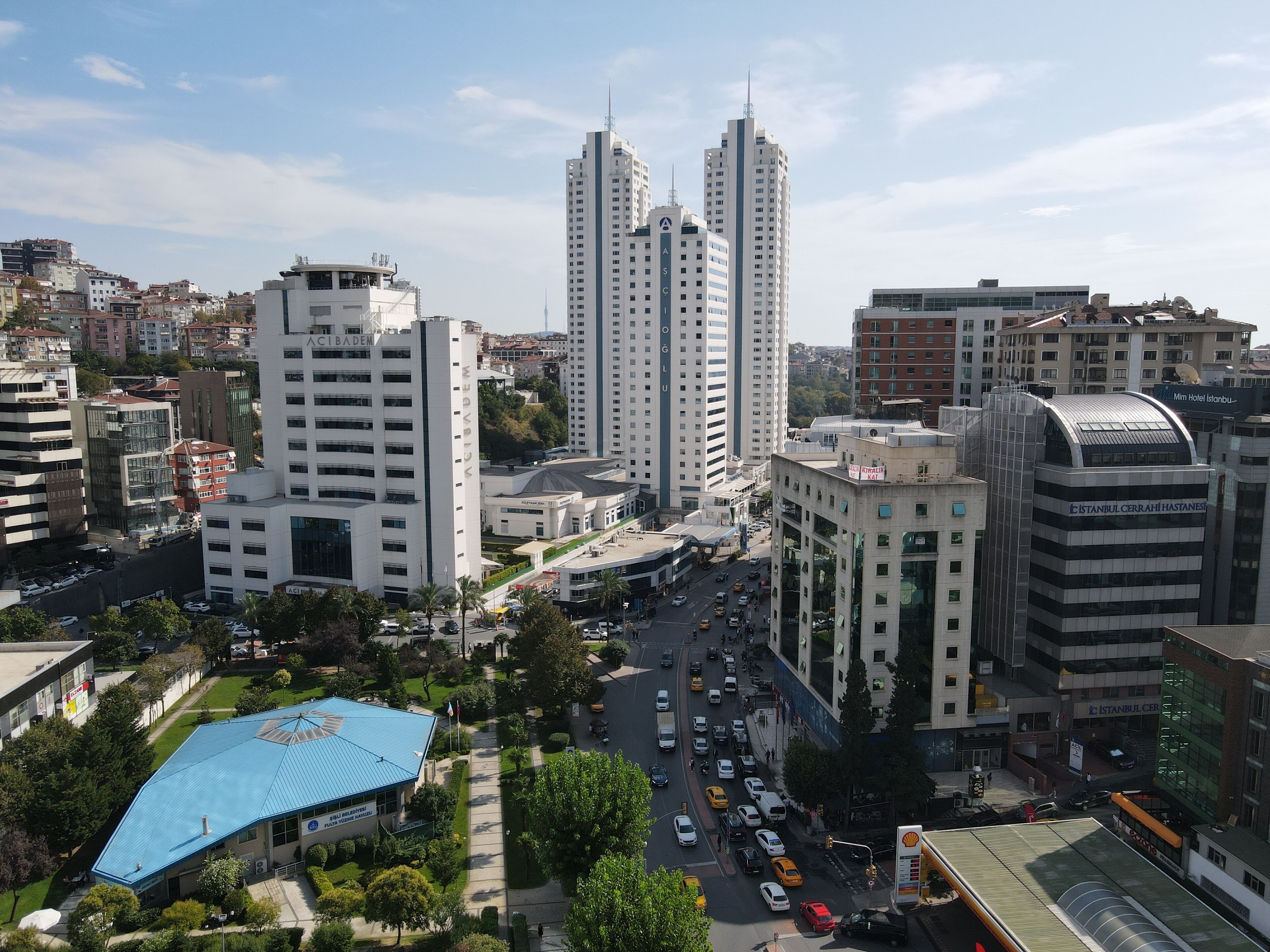 Un paisaje urbano con edificios modernos y altos, una calle con movimiento y un edificio de techo azul con un cartel.