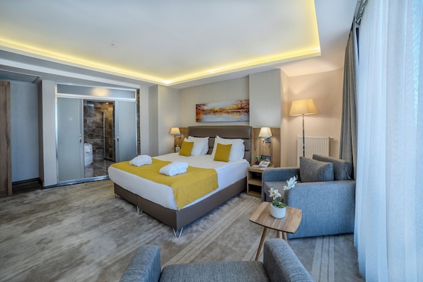A modern hotel room with a large bed, two bedside tables, a seating area with a sofa and armchairs, and a view of the city. - Royal Termal Hotel (Bursa)
