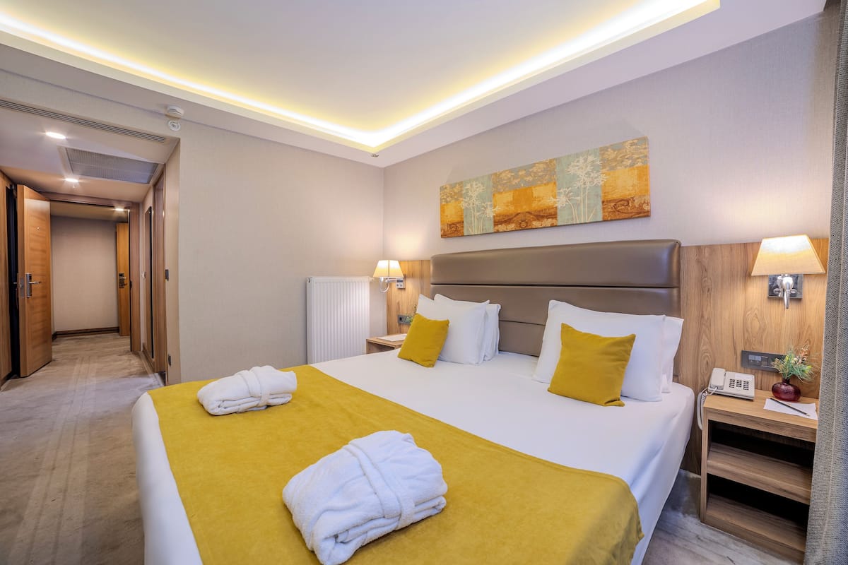 A hotel room with a large bed, a bedside table with a lamp, a painting on the wall, and a hallway leading to another room.