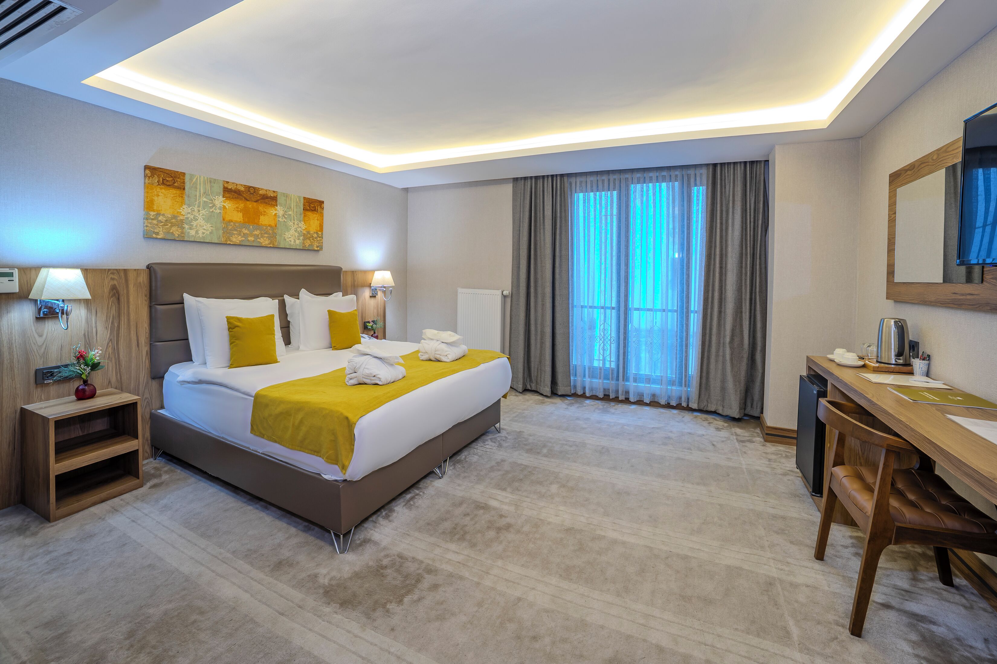 A modern hotel room with a large bed, bedside tables, a desk, and a large window with curtains.