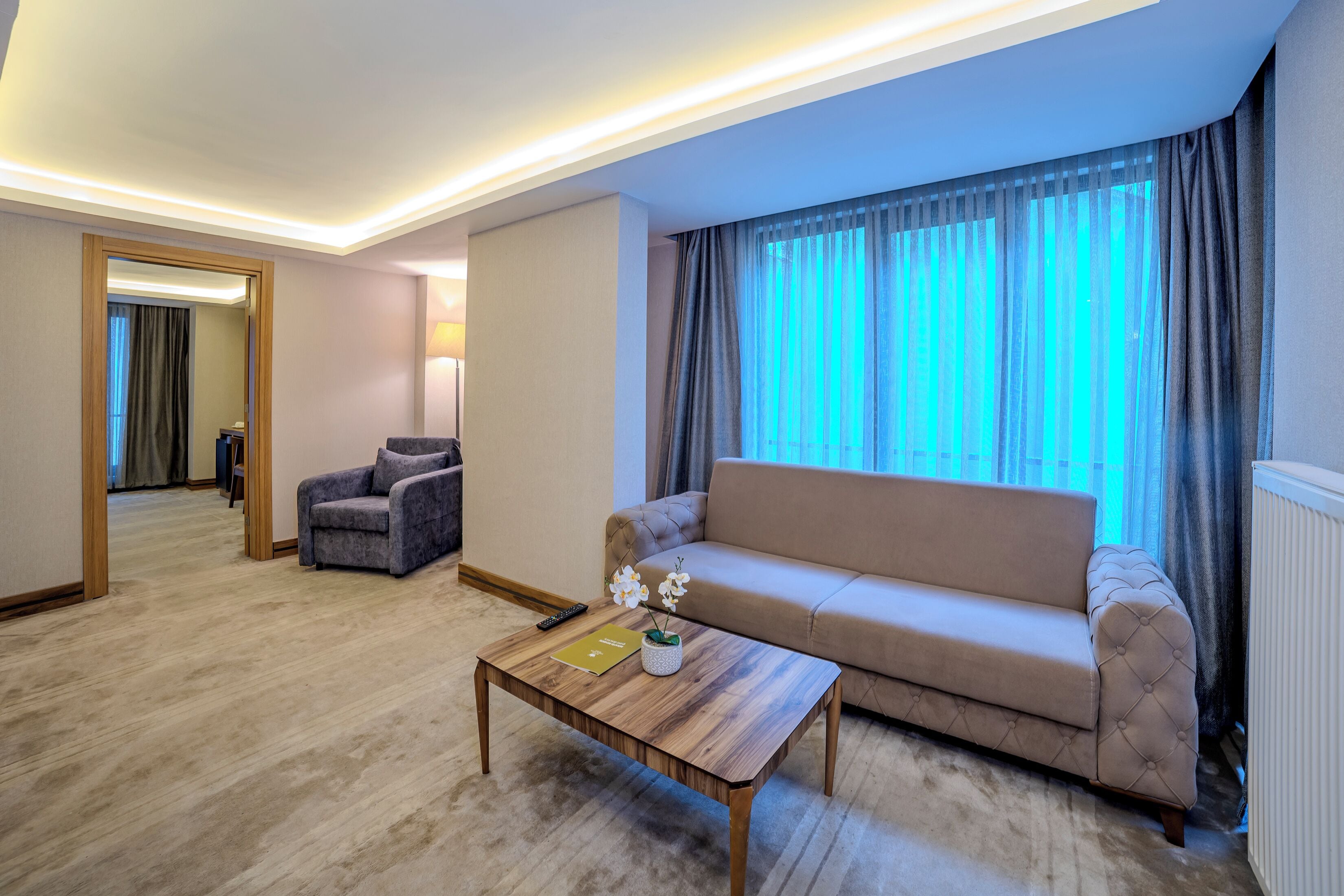 Una sala de estar moderna con sofá, sillón y mesa de centro. Hay un espejo, una lámpara y una ventana con cortinas.