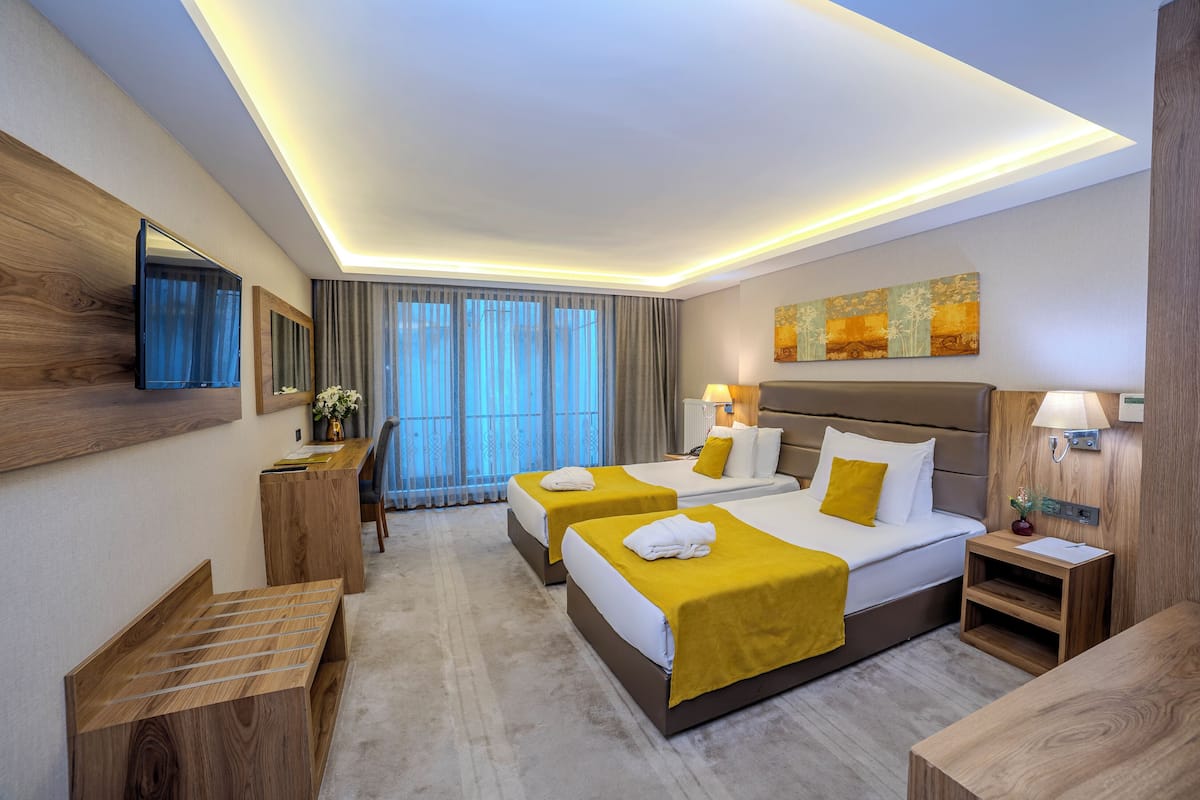 A modern hotel room with two beds, a desk, a TV, and a large window with curtains.