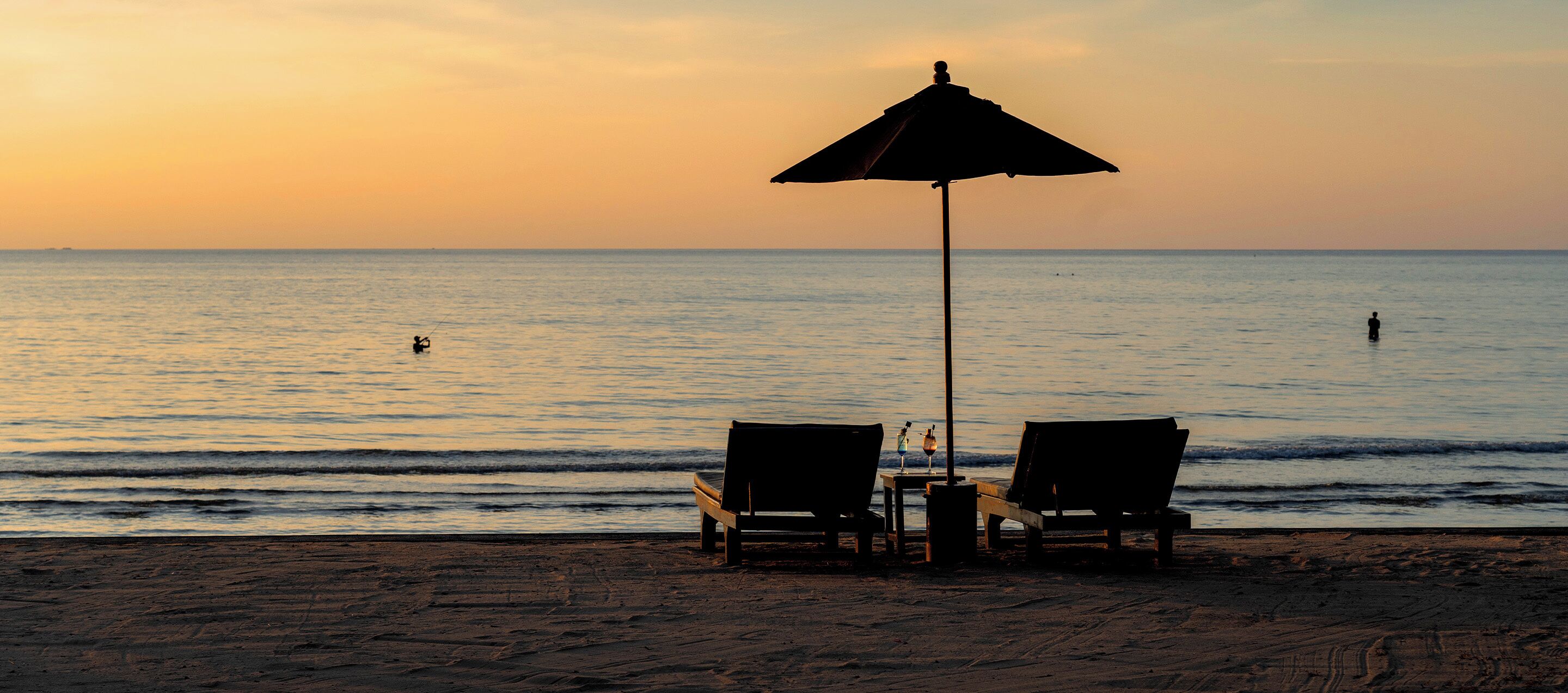 Escena de playa con dos sillas de madera y una mesa bajo un paraguas grande, con vista al mar.
