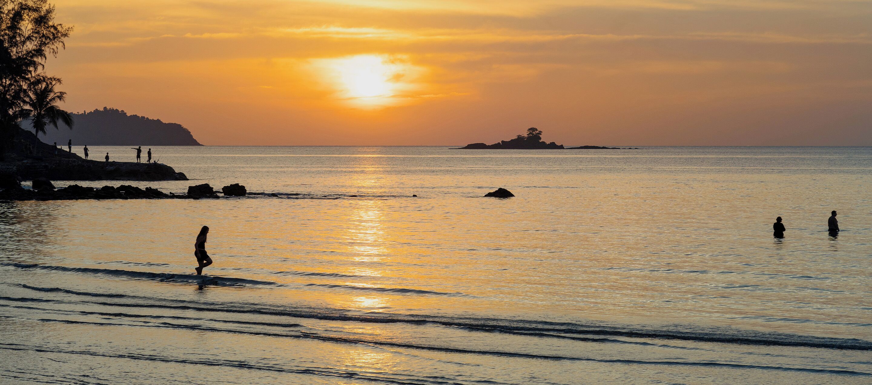 A sunset over a calm sea with rocky shores and a few people in the water.