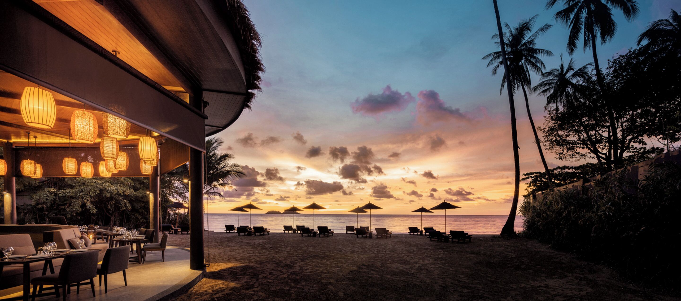 Ein Restaurant direkt am Strand mit Außenbereich und Sonnenschirmen, mit Blick auf den Ozean bei Sonnenuntergang.