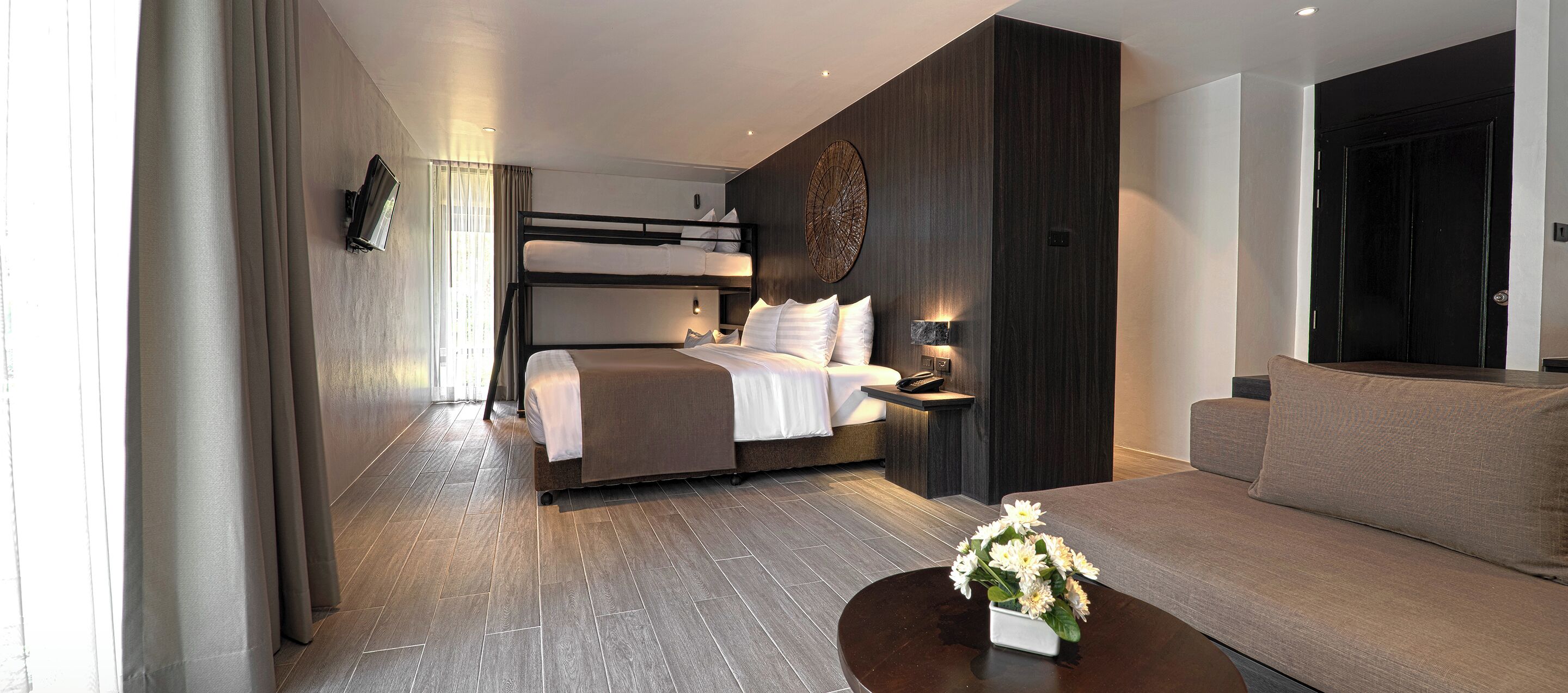 A modern hotel room with a bunk bed, a sofa, a bedside table with a lamp, and a flower vase on a coffee table.