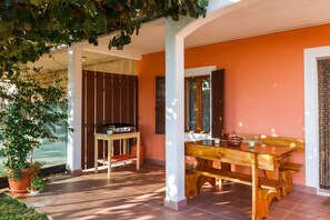 An outdoor patio area with a wooden table and benches, surrounded by orange walls and a covered section with a wooden table and a stove. - Holiday house Ines (Barban)