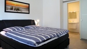 Une chambre à coucher avec un lit, une table de chevet, un tableau encadré au mur et une salle de bain visible à travers une porte ouverte.