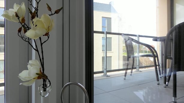Un balcon doté d’une balustrade en verre, d’une chaise et d’un vase contenant des fleurs blanches.