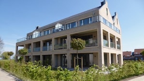 Un immeuble résidentiel à plusieurs étages, doté de balcons, d’une allée pavée et de jeunes arbres au premier plan.