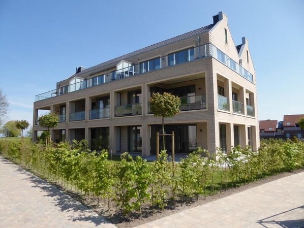 A multi-story residential building with balconies, a paved walkway, and a row of young trees in the foreground.