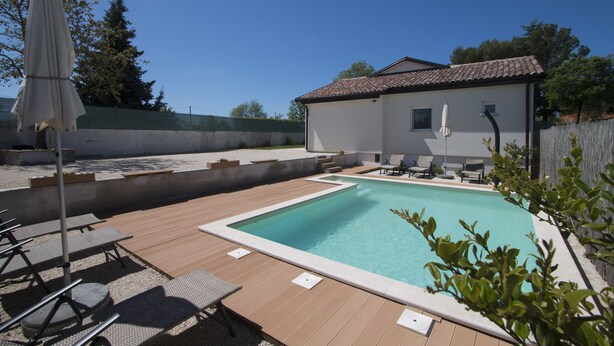 Una piscina con sillas de descanso y una sombrilla cubierta, rodeada por una terraza de madera y un edificio blanco con tejado de tejas.